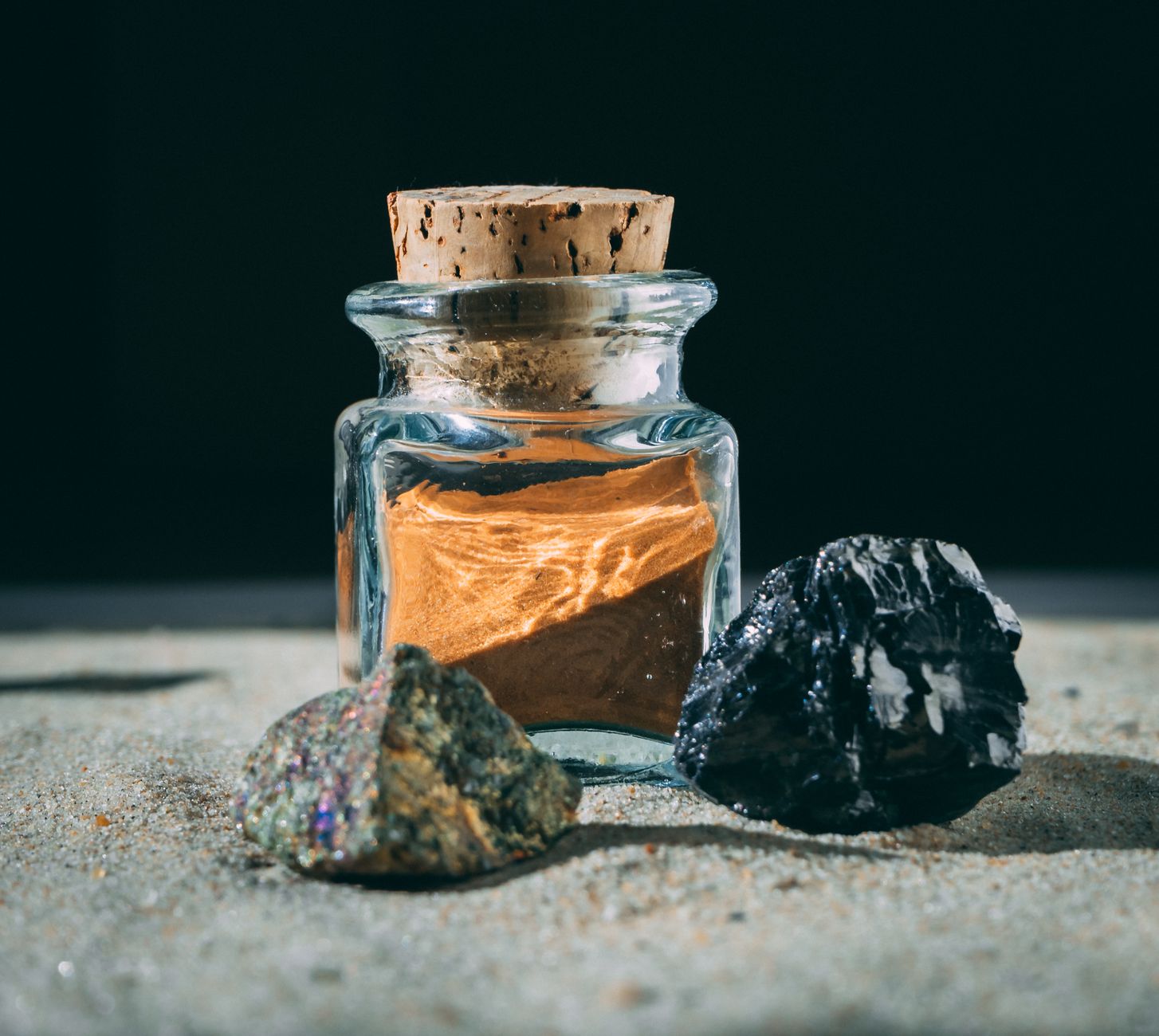 A glass jar with cork lid next to two stones on a sandy surface, under sunlight.