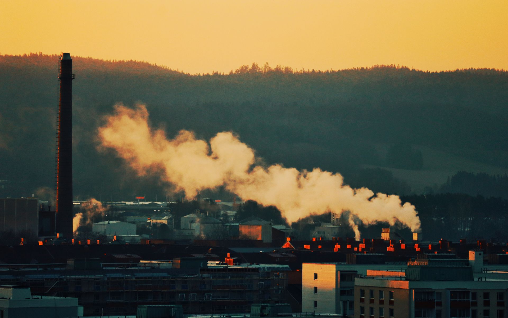 A factory chimney releases smoke into the sunset sky over a townscape surrounded by hills.