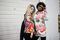 两名时尚女性以现代城市为背景，穿着彩色图案连帽衫，戴着太阳镜