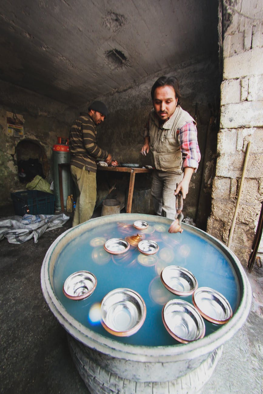 Two artisans skillfully work on metal bowls in a rustic workshop setting.