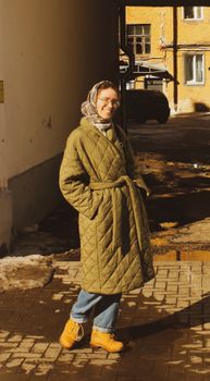 Smiling woman wearing a quilted coat and headscarf enjoys a sunny day outdoors.