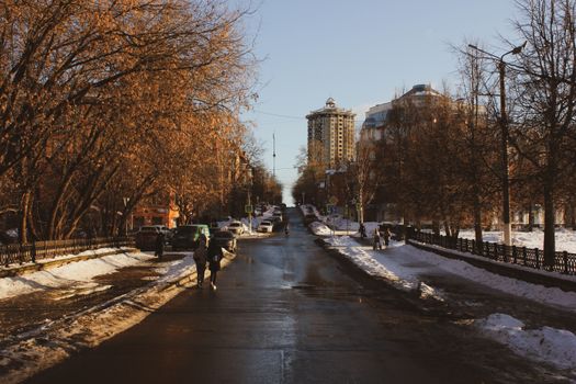 A winter street scene showing people walking along a snowy road lined with trees and buildings.