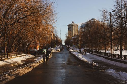 Free A winter street scene showing people walking along a snowy road lined with trees and buildings. Stock Photo