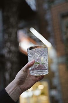 A hand raises a mojito in a crystal glass, toasting outdoors in a lively setting.