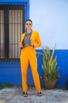 Stylish woman in orange suit and sunglasses stands confidently by a colorful wall.