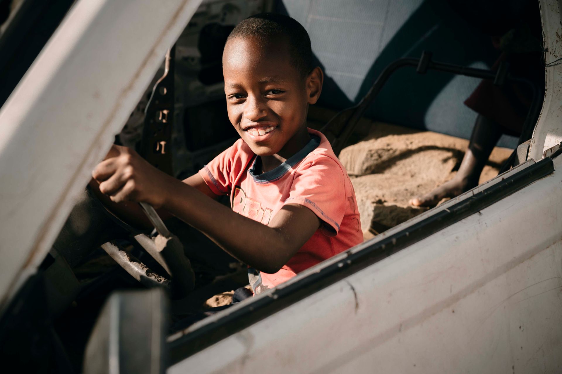 A cheerful young boy smiling while seated in the driver's seat of a car on a sunny day.