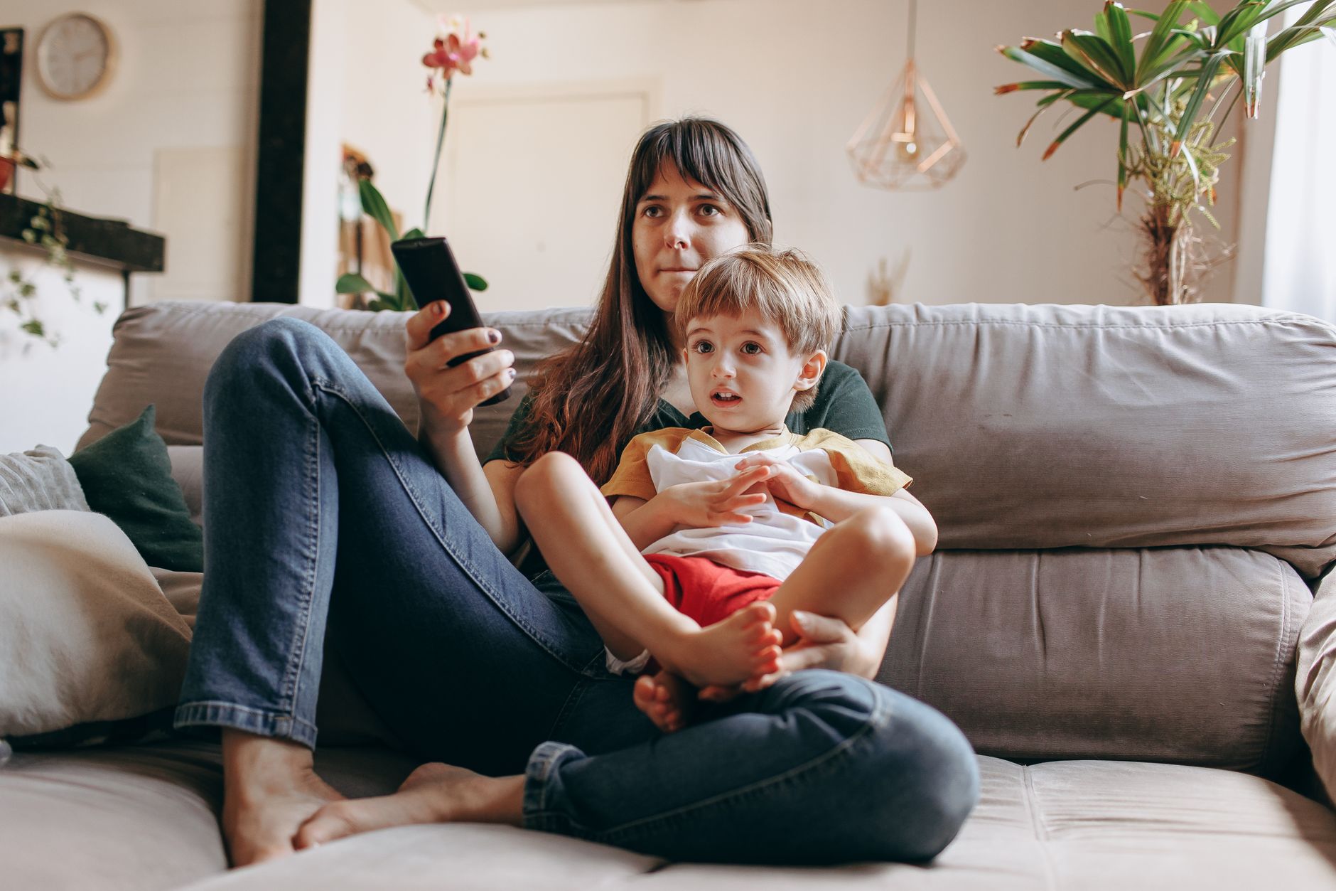 A mother and son sitting on the couch watching TV together in a cozy living room.