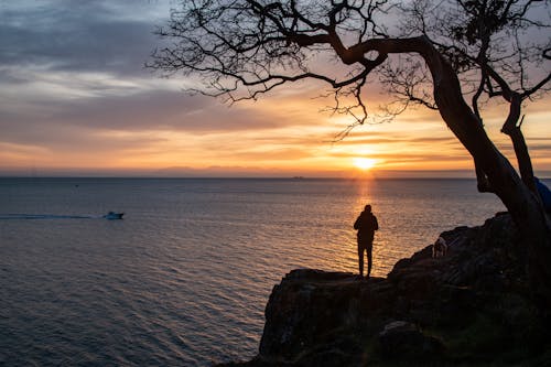 Free Silhouette of Person Standing on Rock Formation Near Body of Water during Sunset Stock Photo