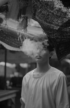 A monochrome image of a man standing outdoors with smoke obscuring his face, creating a mysterious vibe.