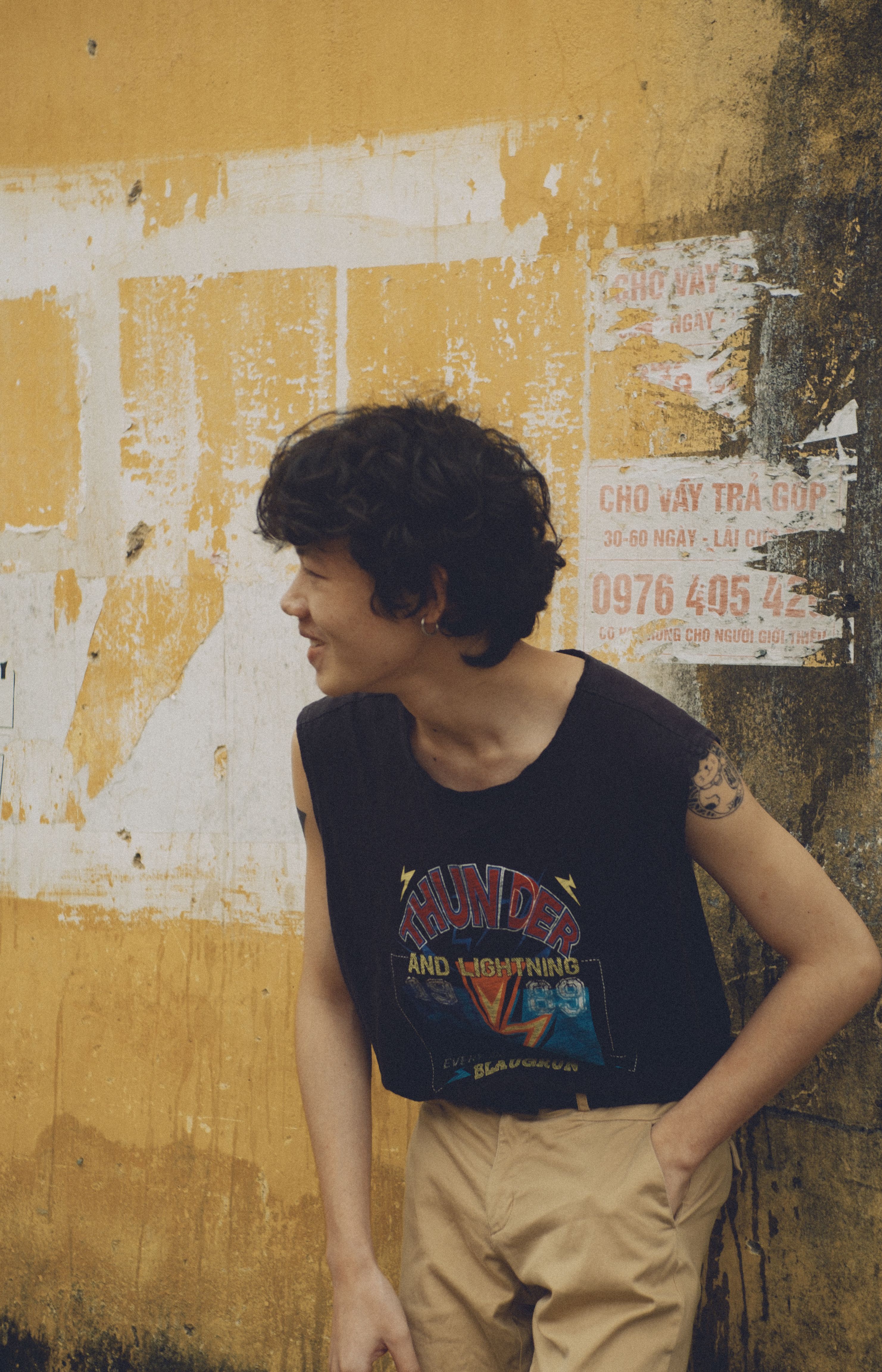 Young man with curly hair leaning and smiling against a rustic yellow wall.