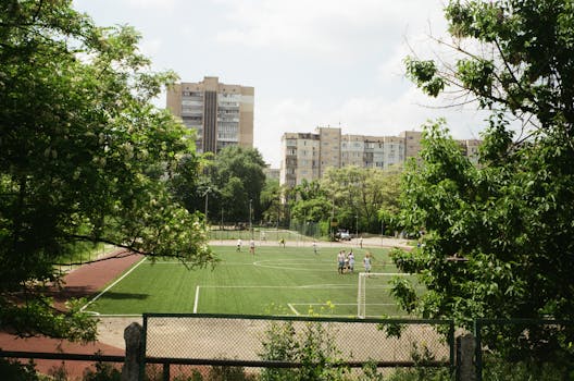 Aerial view of an urban soccer field with players during a match surrounded by greenery and residential buildings.