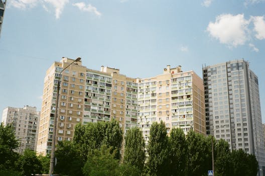Residential high-rise buildings in Kyiv with green trees beneath a clear blue sky.