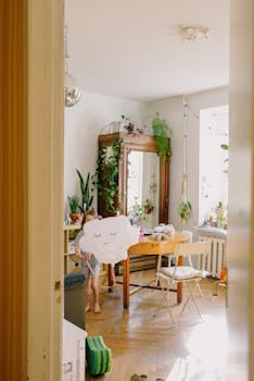 Child playing with cloud-shaped pillow in a sunny, plant-filled interior with wooden floors.