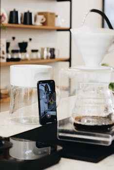 A sleek kitchen scene featuring a smartphone recording drip coffee brewed in a glass container.