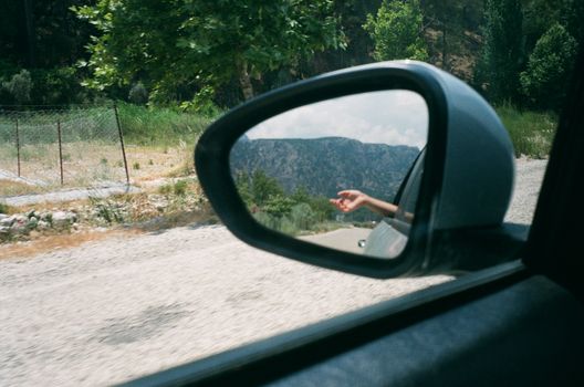 Side mirror view of road trip scenery with a hand outside the car window.