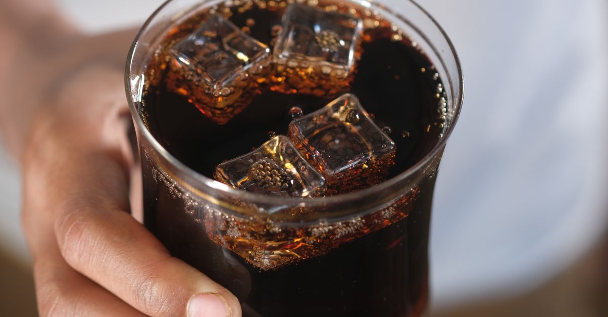Close-up de uma mão segurando um copo de refrigerante escuro com cubos de gelo, proporcionando uma sensação refrescante.