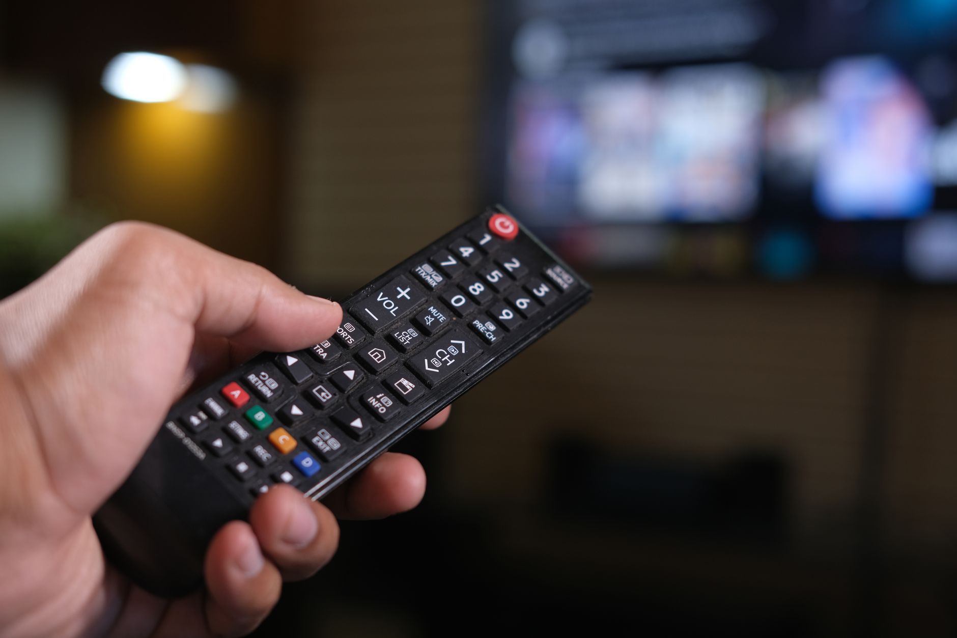 Person holding a remote control with a blurred TV screen in the background.