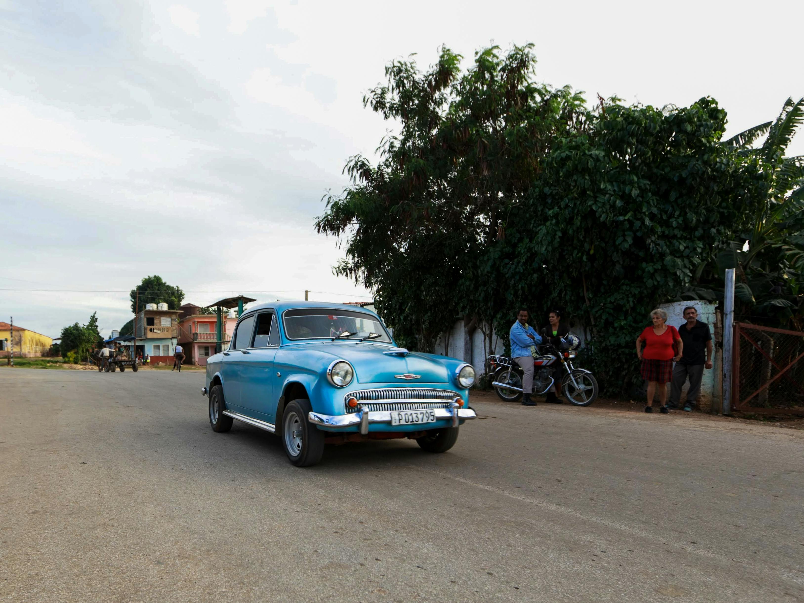 A retro blue car drives on a suburban street, capturing classic automotive charm.