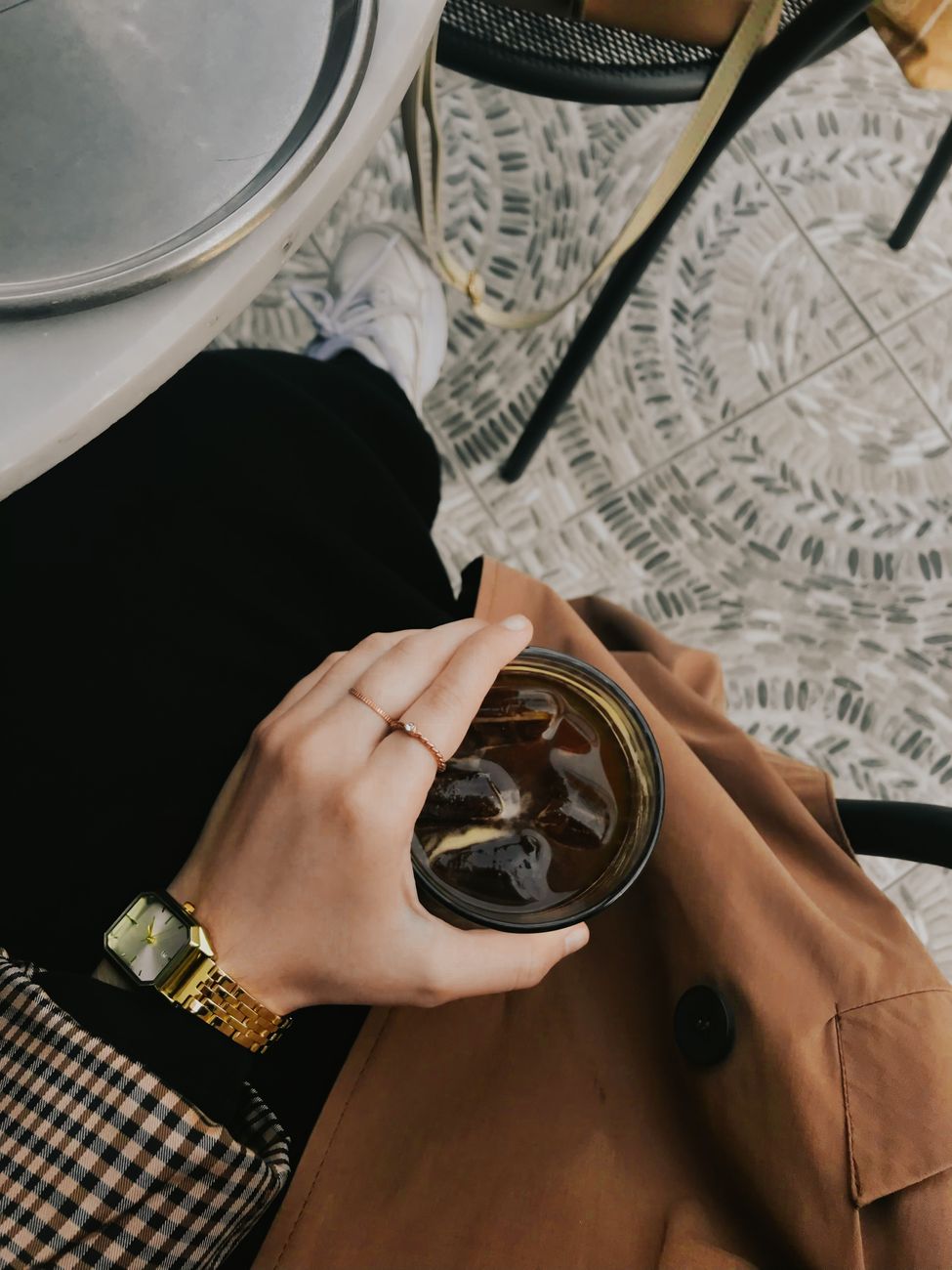 A fashionable individual holding an iced coffee at a chic outdoor café, featuring a gold watch and autumn attire.