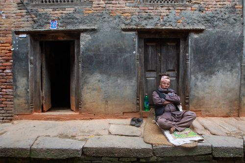 Free An elderly man in traditional wear sits cross-legged outside a historic building. Stock Photo