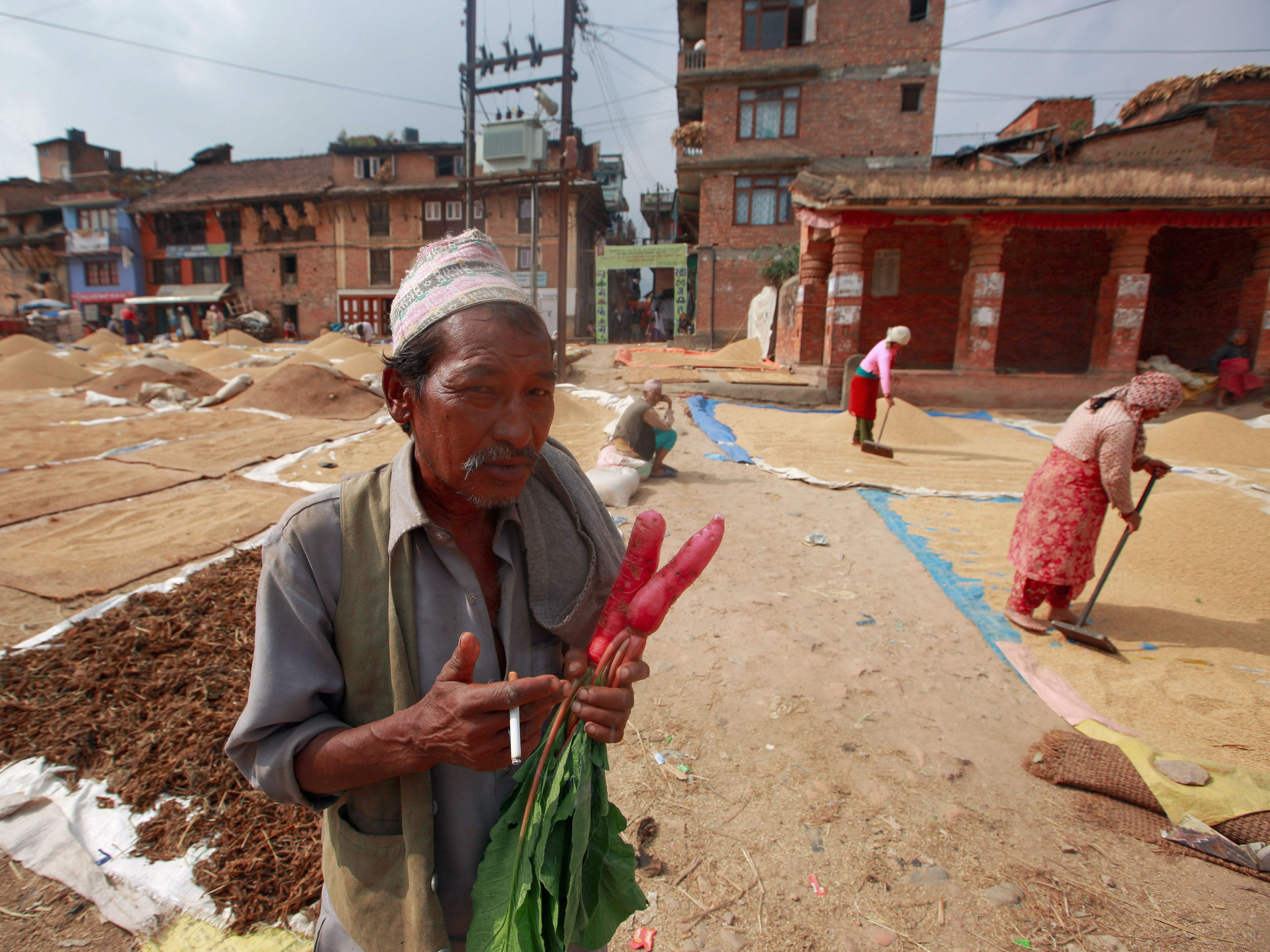 A native farmer stands outdoors holding fresh radishes with people working in the background.