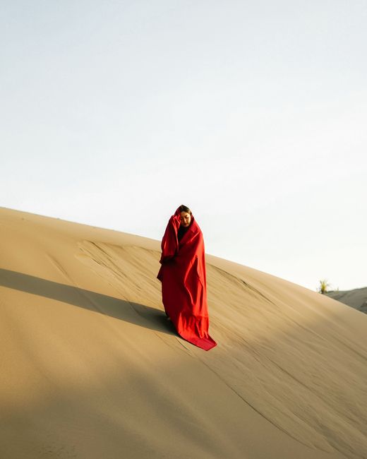 A person stands wrapped in a red blanket on a desert sand dune in Yogyakarta, Indonesia.