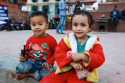 Two adorable children sitting on a city street, wearing colorful clothes and smiling playfully.