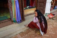 Woman with long hair sits on steps by shop displaying colorful scarves, reflecting culture.