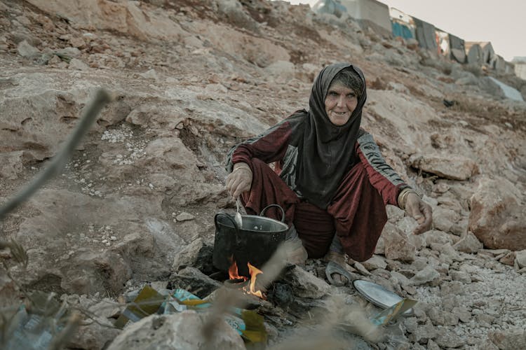 Elderly Woman Cooking While Sitting On Rocky Ground