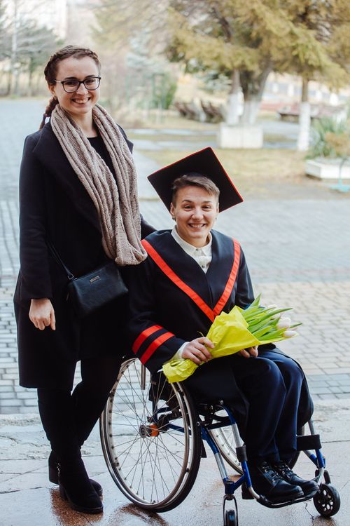 Free Two adults celebrate graduation outdoors, one in a wheelchair, holding flowers. Stock Photo