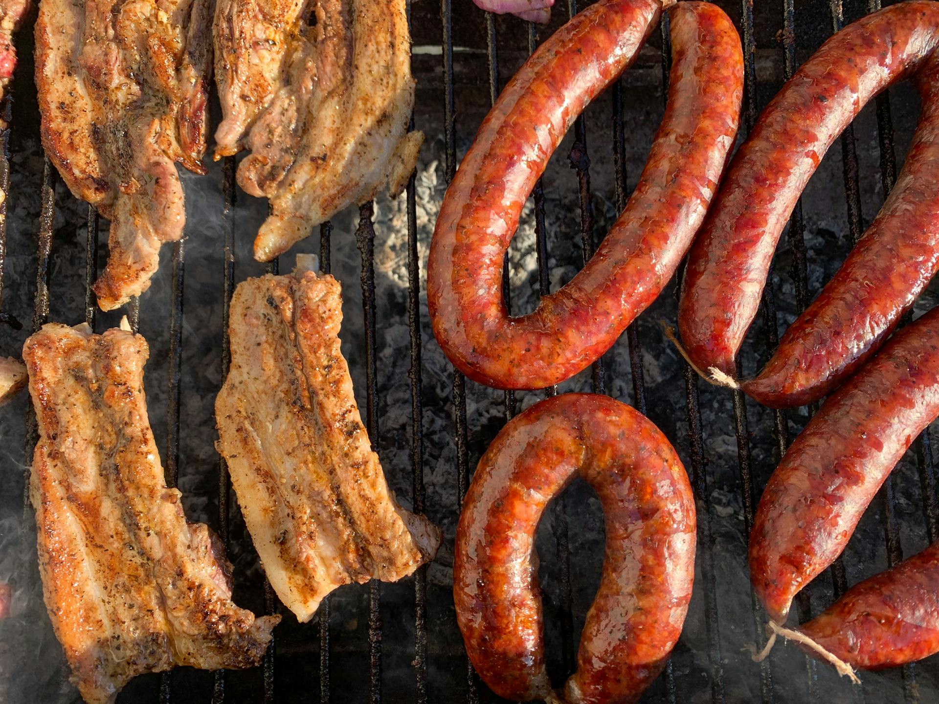 Sizzling sausages and ribs on a hot charcoal grill, emitting savory smoke.