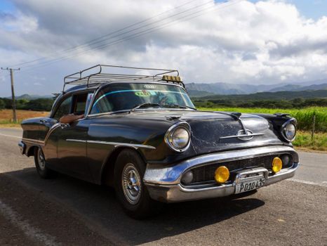 A beautifully preserved vintage car travels on a picturesque country road under a bright blue sky.