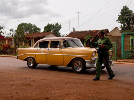 Classic yellow car parked on a rustic street with trees and houses in the background.