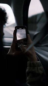 A person taking a photo through a car window using a smartphone, showcasing a moody landscape.