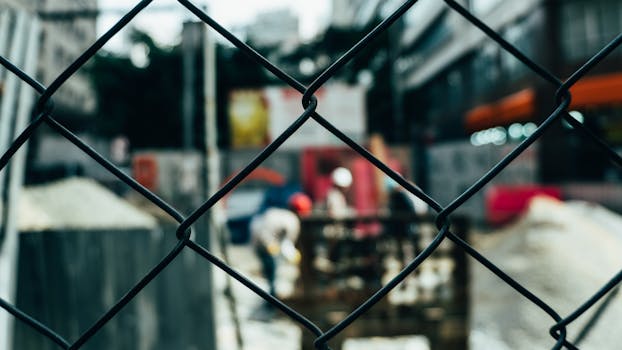 Blurred view of a construction site seen through a chain link fence, depicting industrial activity.