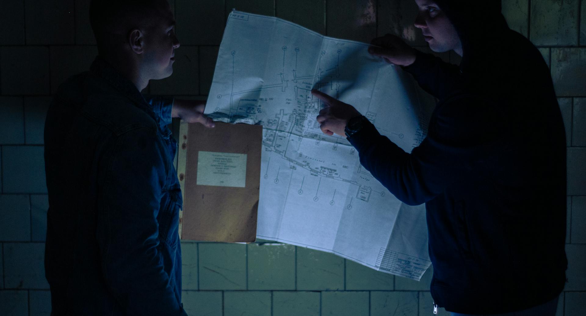 Two individuals reviewing a detailed blueprint in a low-light setting, focusing on planning and collaboration.