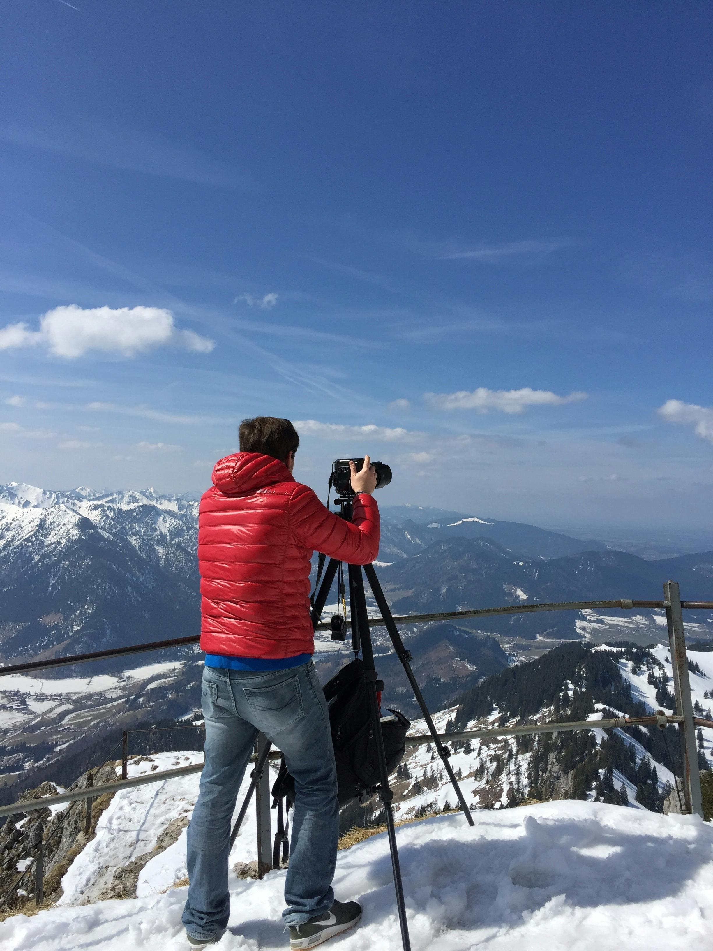 Photographer in a red jacket captures snowy Bavarian Alps with tripod.