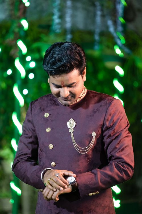 Free South Asian man in a traditional maroon suit adjusting his wristwatch with a lush green backdrop. Stock Photo