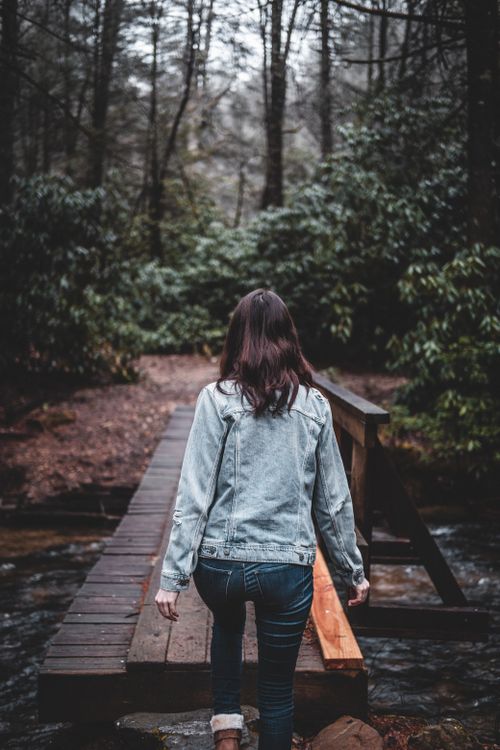 Free A woman in a denim jacket walks across a wooden footbridge in a lush forest, evoking a sense of adventure. Stock Photo