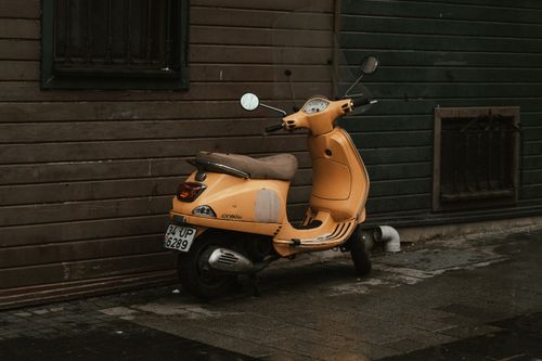Free A vibrant yellow scooter leans against a rustic wooden wall on a damp pavement. Stock Photo