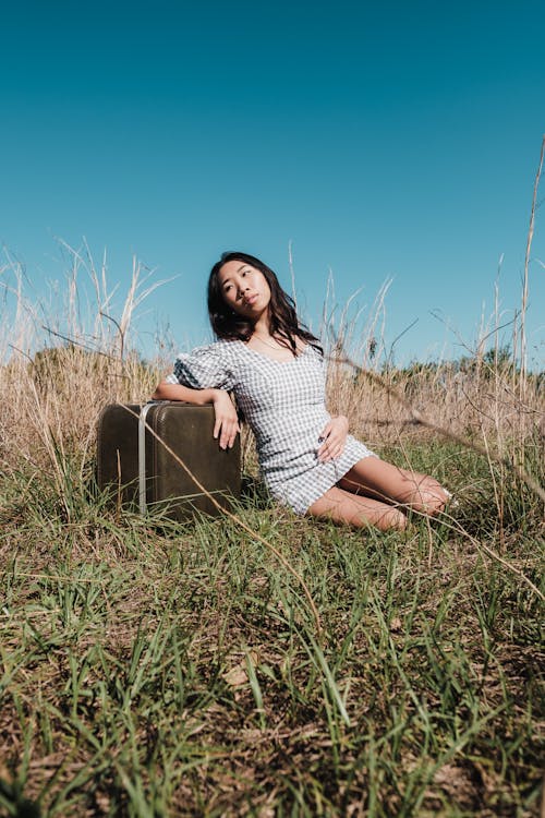 Woman in Plaid Mini Dress Sitting and Leaning on a Suitcase on Grass Field