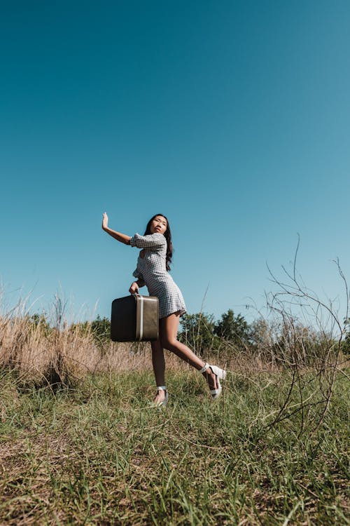 Woman Posing in Plaid Mini Dress Holding a Suitcase on a Grass Field