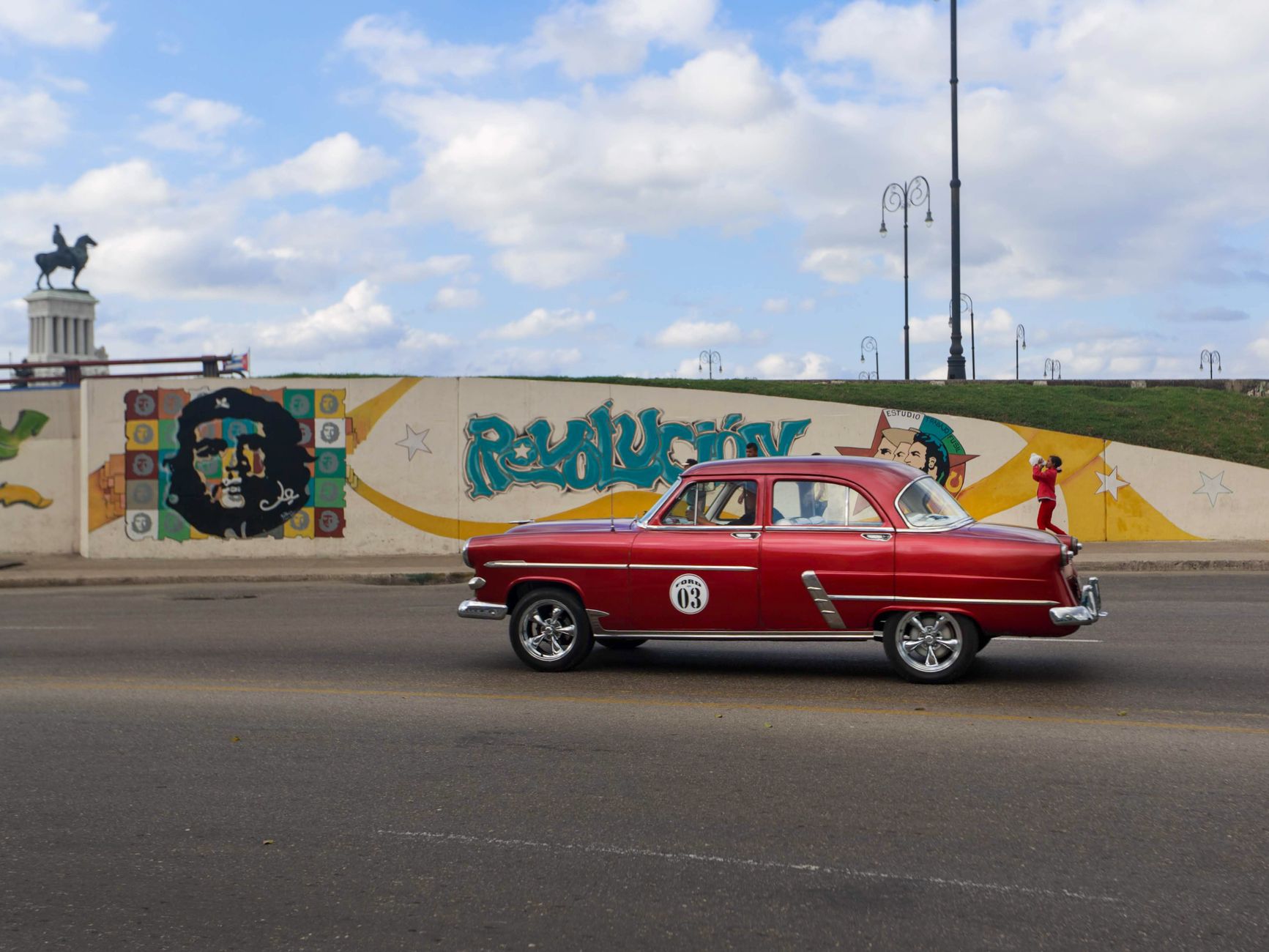Vintage red car cruising by Cuban street art in Havana, featuring Che Guevara mural.