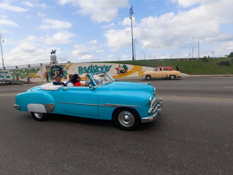 A classic blue convertible car in Havana with vibrant street art and people riding.