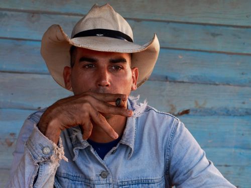 Free Close-up of a man wearing a cowboy hat, smoking a cigar against a rustic wood background. Stock Photo