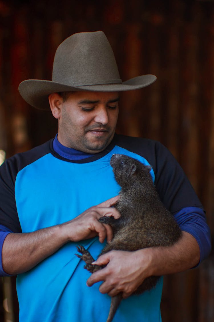 A Man Wearing A Brown Cowboy Hat Holding A Black Rodent