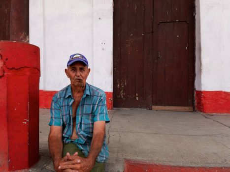 Elderly man with cap sitting beside a rustic doorway on a sunny day.