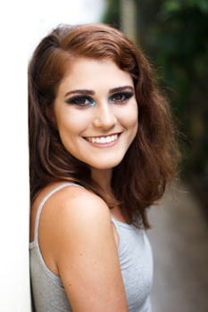 Portrait of a young woman with glamorous makeup and a bright smile in a casual outdoor setting.