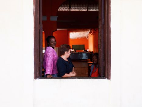 Three women chatting and enjoying leisure time by an open window, indoors with warm lighting.
