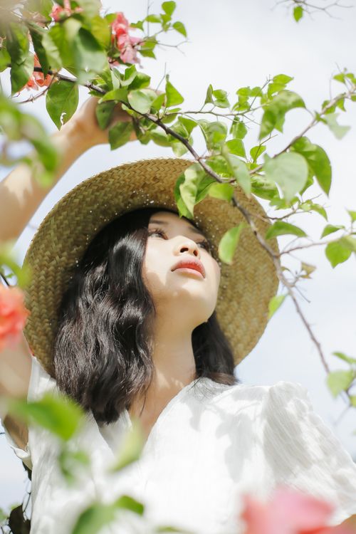 Free A young woman wearing a straw hat looks up amidst lush green leaves and flowers. Stock Photo
